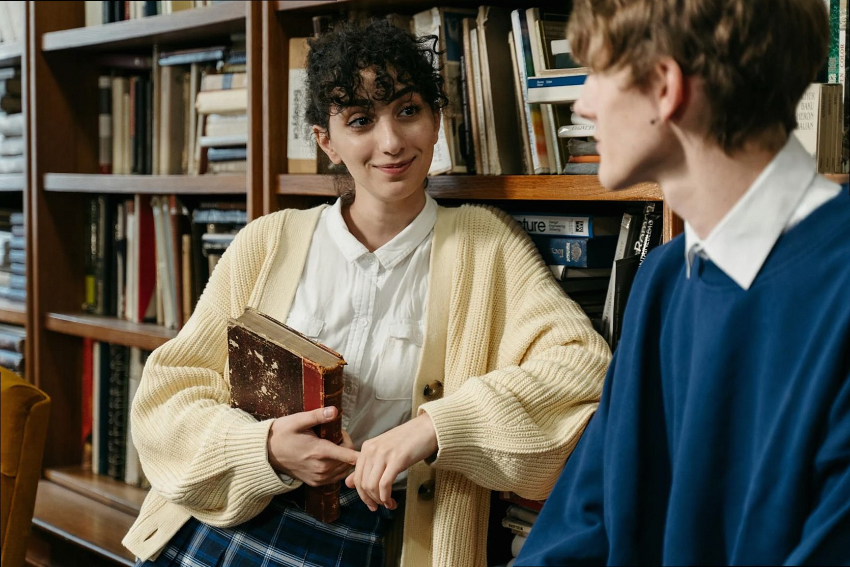 Dos estudiantes mantienen una alegre conversación en una biblioteca con estanterías de fondo.